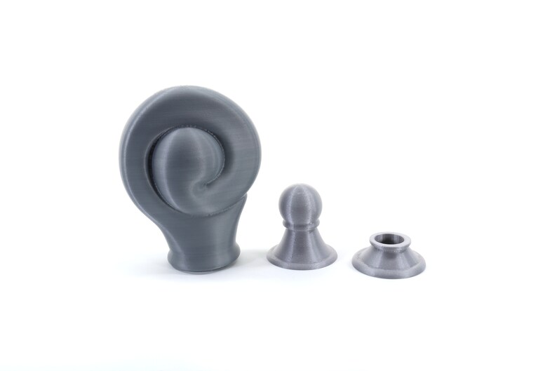 K&ouml;nnte beinhalten: Drei graue 3D-gedruckte Objekte auf wei&szlig;em Hintergrund. Das gr&ouml;&szlig;te Objekt hat ein geschwungenes, spiralf&ouml;rmiges Design. Zwei kleinere Objekte &auml;hneln Schachfiguren, eines mit einem kugelf&ouml;rmigen Kopf und das andere mit einer kreisf&ouml;rmigen &Ouml;ffnung.