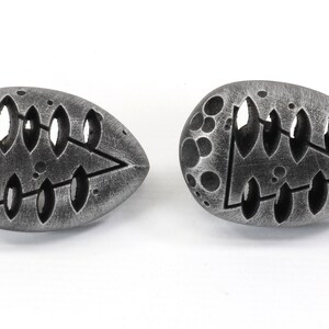May include: Two silver-colored, egg-shaped earrings with a tree design cut out. The earrings have a textured surface with small circles.