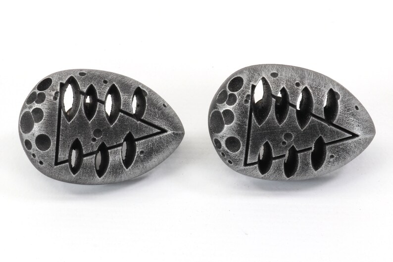 May include: Two silver-colored, egg-shaped earrings with a tree design cut out. The earrings have a textured surface with small circles.