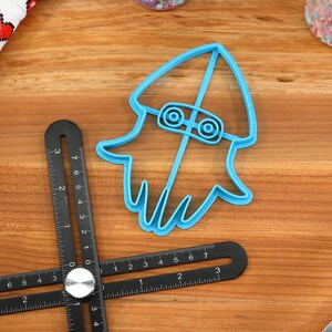 May include: Blue plastic cookie cutter shaped like a squid wearing a hat. The cutter is on a wooden surface with a ruler and a rolling pin.