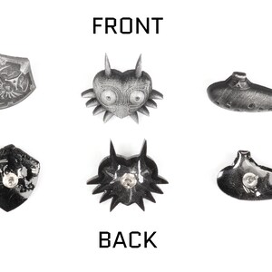 May include: Six silver-coloured metal pins featuring different designs from the Legend of Zelda video game series. The pins are arranged in two rows of three. The top row features a shield, a mask, and an ocarina. The bottom row features a shield, a mask, and an ocarina. The pins are all approximately 5 cm in diameter.
