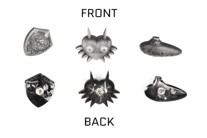 May include: Six silver-coloured metal pins featuring different designs from the Legend of Zelda video game series. The pins are arranged in two rows of three. The top row features a shield, a mask, and an ocarina. The bottom row features a shield, a mask, and an ocarina. The pins are all approximately 5 cm in diameter.