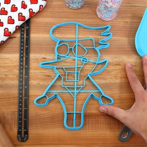 May include: Blue plastic cookie cutter shaped like a cartoon character wearing glasses and a hat. The cutter is on a wooden surface with a rolling pin, a measuring tape, and a spatula.