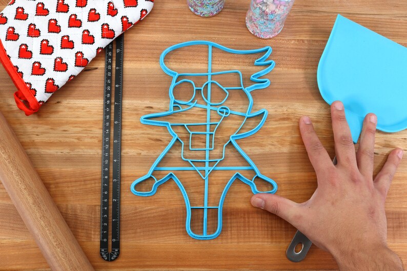 May include: Blue plastic cookie cutter shaped like a cartoon character wearing glasses and a hat. The cutter is on a wooden surface with a rolling pin, a measuring tape, and a spatula.