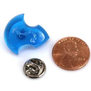 May include: A blue plastic pin in the shape of a crescent moon with a white pin back. The pin is about the size of a US penny.