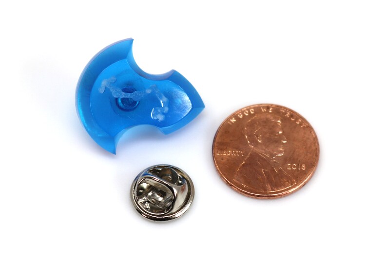 May include: A blue plastic pin in the shape of a crescent moon with a white pin back. The pin is about the size of a US penny.