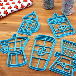 May include: A set of five blue plastic cookie cutters shaped like different coffee brewing methods: a French press, a coffee maker, a coffee cup, an espresso pot, and a pour-over coffee maker.