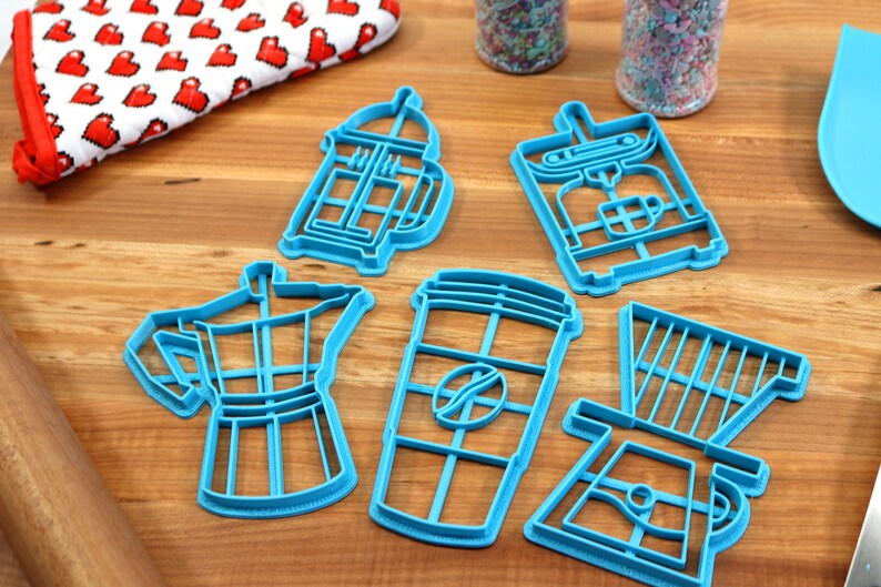 May include: A set of five blue plastic cookie cutters shaped like different coffee brewing methods: a French press, a coffee maker, a coffee cup, an espresso pot, and a pour-over coffee maker.