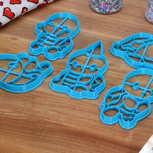May include: Five blue plastic cookie cutters shaped like cartoon characters. The cutters are on a wooden surface.