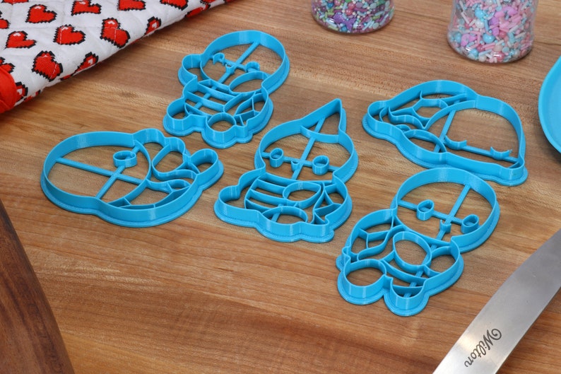 May include: Five blue plastic cookie cutters shaped like cartoon characters. The cutters are on a wooden surface.