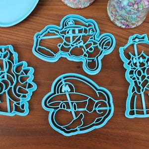 May include: Set of five blue plastic cookie cutters shaped like characters from the Super Mario Bros. video game franchise. The cutters include Mario, Luigi, Princess Peach, Bowser, and a Goomba.