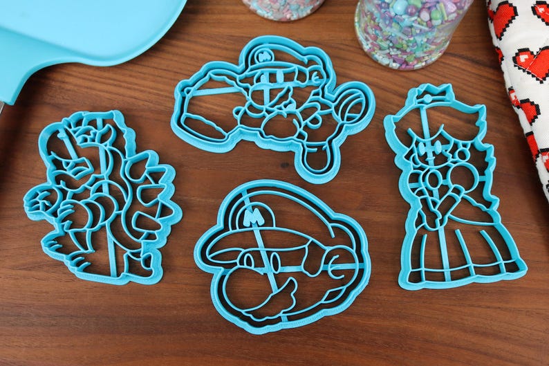 May include: Set of five blue plastic cookie cutters shaped like characters from the Super Mario Bros. video game franchise. The cutters include Mario, Luigi, Princess Peach, Bowser, and a Goomba.