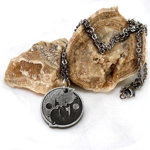 May include: A silver chain necklace with a round pendant featuring a yin yang symbol with two stylized figures inside. The pendant is made of metal and has a black and silver finish.