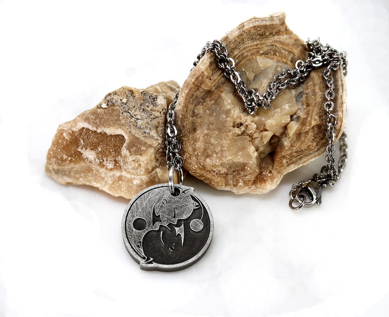 May include: A silver chain necklace with a round pendant featuring a yin yang symbol with two stylized figures inside. The pendant is made of metal and has a black and silver finish.