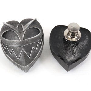 May include: A silver heart-shaped pin with a grinning face and sharp teeth. The back of the pin is black and has a metal clasp.