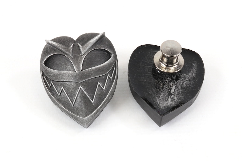 May include: A silver heart-shaped pin with a grinning face and sharp teeth. The back of the pin is black and has a metal clasp.