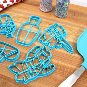 May include: A set of five blue plastic cookie cutters shaped like a bottle, a cat, a snowman, and two other characters. The cutters are on a wooden surface with a blue spatula.