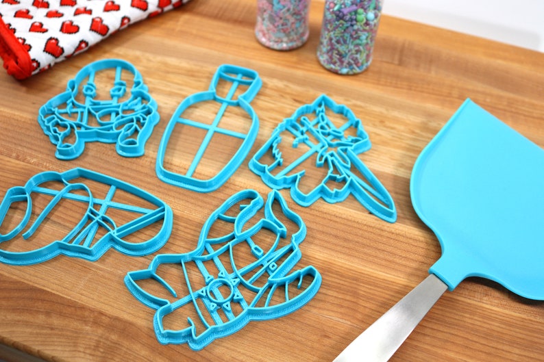 May include: A set of five blue plastic cookie cutters shaped like a bottle, a cat, a snowman, and two other characters. The cutters are on a wooden surface with a blue spatula.