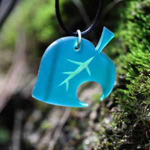May include: A teal blue leaf-shaped pendant with a white silhouette of a leaf inside. The pendant is hanging from a black cord.
