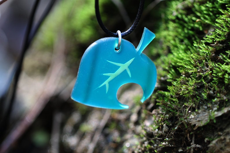 May include: A teal blue leaf-shaped pendant with a white silhouette of a leaf inside. The pendant is hanging from a black cord.