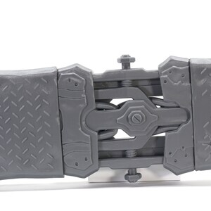 May include: A close-up of a gray plastic toy with a detailed, articulated design. The toy has a series of interlocking pieces that connect with screws. The toy is made of a textured plastic with a diamond pattern.
