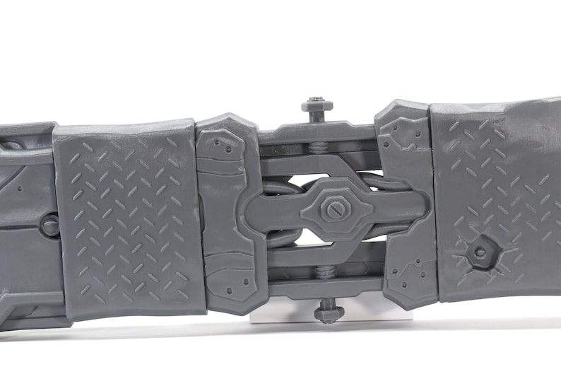 May include: A close-up of a gray plastic toy with a detailed, articulated design. The toy has a series of interlocking pieces that connect with screws. The toy is made of a textured plastic with a diamond pattern.