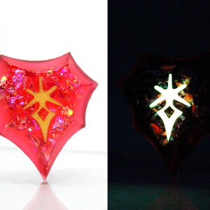 May include: A red, heart-shaped resin pendant with a gold-colored symbol inside. The pendant glows in the dark, revealing a white symbol.