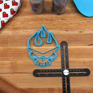May include: Blue plastic cookie cutter shaped like a skull with flames on top. The cutter is on a wooden surface with a ruler and a rolling pin.