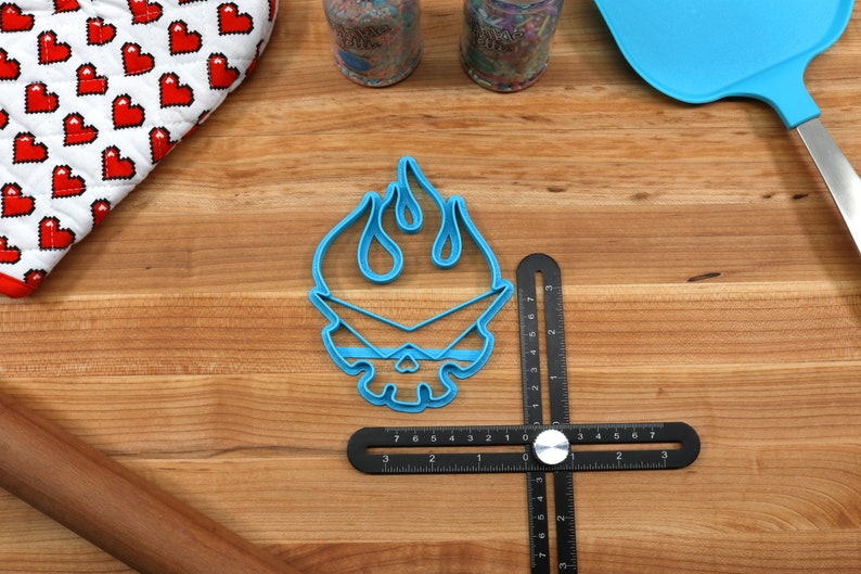 May include: Blue plastic cookie cutter shaped like a skull with flames on top. The cutter is on a wooden surface with a ruler and a rolling pin.