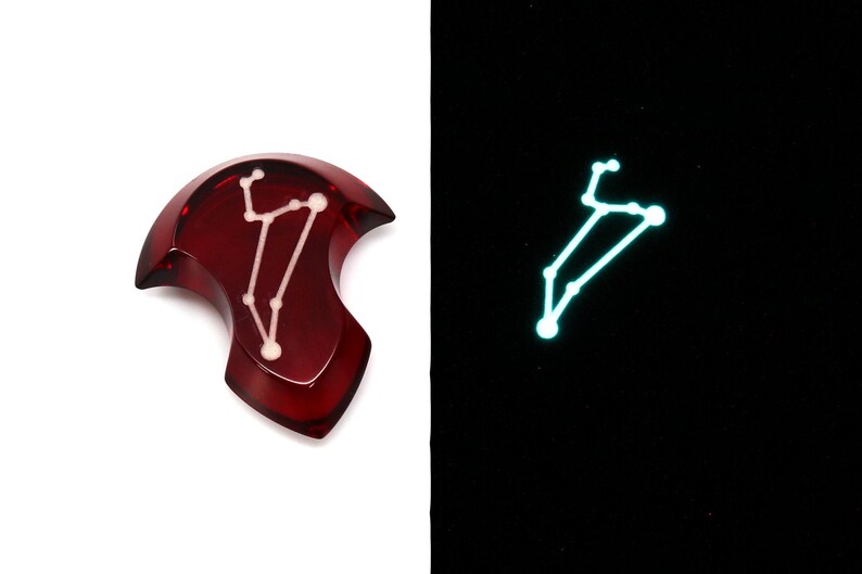 May include: A red resin pendant with a white constellation design that glows in the dark. The pendant is shaped like a crescent moon.