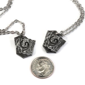 May include: Two silver necklaces with matching pendants. Each pendant is a shield-shaped piece of metal with a textured surface and a stylized letter 'G' engraved on it. A US dime is included for scale.