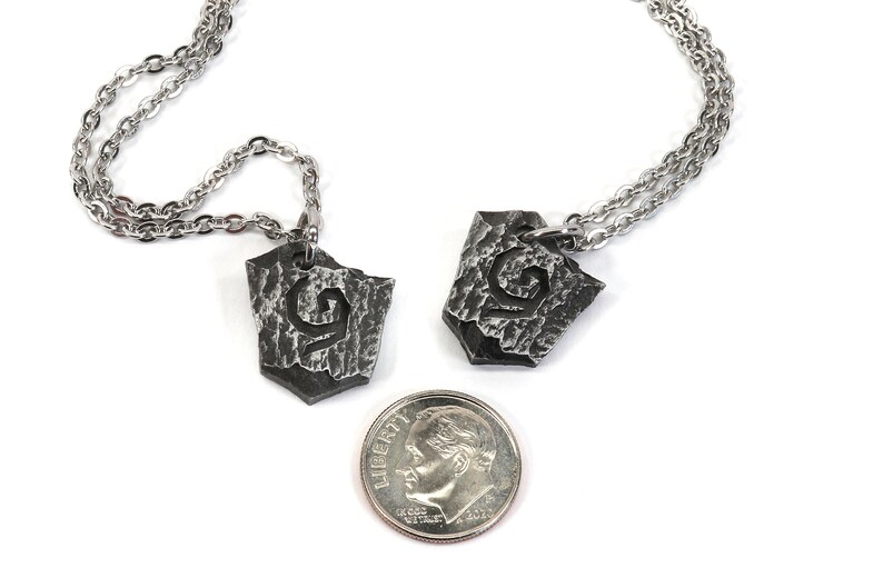 May include: Two silver necklaces with matching pendants. Each pendant is a shield-shaped piece of metal with a textured surface and a stylized letter 'G' engraved on it. A US dime is included for scale.