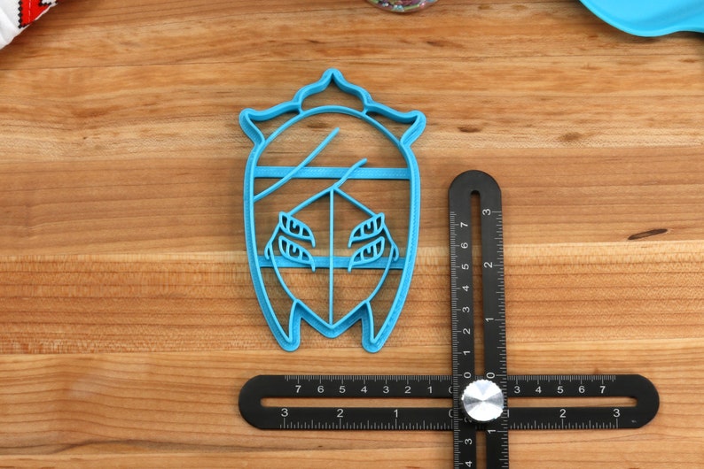 May include: A blue plastic cookie cutter shaped like a stylized face with a crown on top. The cutter is about 2 inches tall and is sitting on a wooden surface.