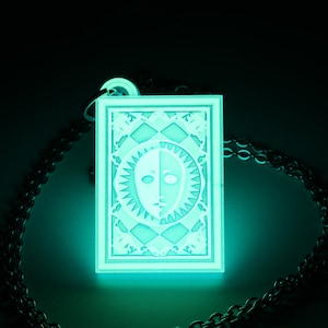 Puede incluir: Un collar con un colgante verde fluorescente con un dise&ntilde;o blanco de una cara con un sol y una luna a cada lado. El colgante est&aacute; en un marco cuadrado con un borde decorativo.