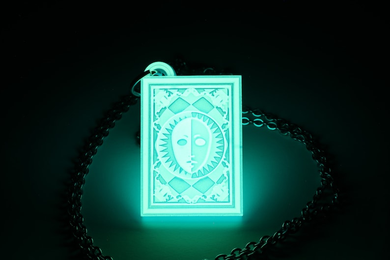 Puede incluir: Un collar con un colgante verde fluorescente con un dise&ntilde;o blanco de una cara con un sol y una luna a cada lado. El colgante est&aacute; en un marco cuadrado con un borde decorativo.