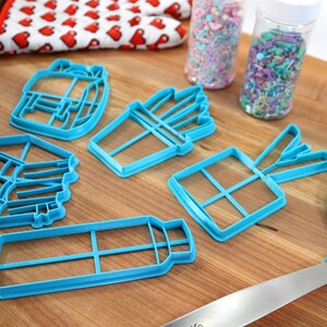 K&ouml;nnte beinhalten: Ein Satz von f&uuml;nf blauen Plastik-Ausstechern in Form einer Geschenkbox, einer Pommes-Frites-Verpackung, einer Wasserflasche, eines Blumenstrau&szlig;es und eines Stapels B&uuml;cher. Die Ausstecher befinden sich auf einer Holzoberfl&auml;che.