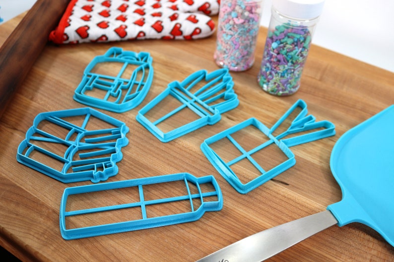 K&ouml;nnte beinhalten: Ein Satz von f&uuml;nf blauen Plastik-Ausstechern in Form einer Geschenkbox, einer Pommes-Frites-Verpackung, einer Wasserflasche, eines Blumenstrau&szlig;es und eines Stapels B&uuml;cher. Die Ausstecher befinden sich auf einer Holzoberfl&auml;che.