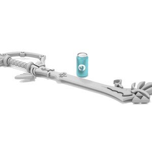 K&ouml;nnte beinhalten: Eine wei&szlig;e Plastik-Replik der Kingdom Key, ein schl&uuml;sself&ouml;rmiges Schwert aus der Kingdom Hearts-Videospielserie. Das Schwert liegt auf einer wei&szlig;en Oberfl&auml;che neben einer blauen Dose.