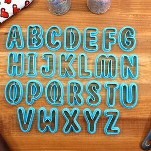 May include: A set of 26 blue plastic alphabet cookie cutters on a wooden surface. The cutters are in uppercase and are arranged in alphabetical order.
