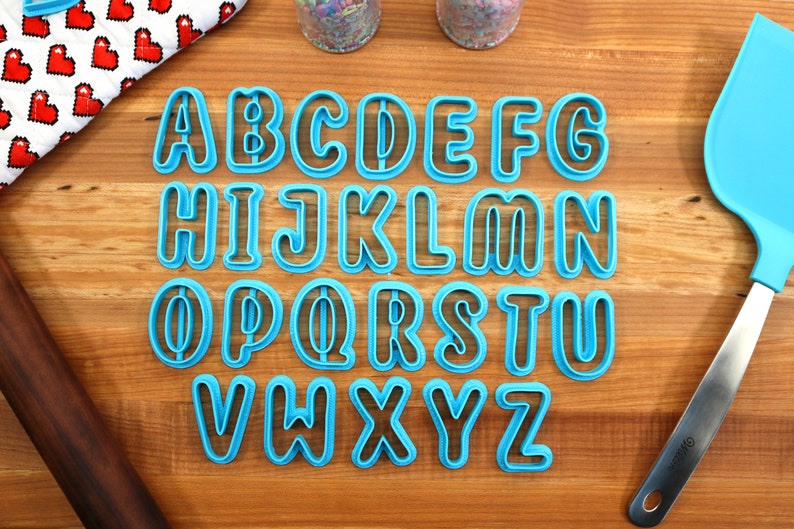 May include: A set of 26 blue plastic alphabet cookie cutters on a wooden surface. The cutters are in uppercase and are arranged in alphabetical order.
