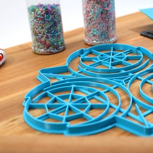 May include: Blue plastic cookie cutters shaped like a snowflake with a circular design. The cutters are on a wooden surface.
