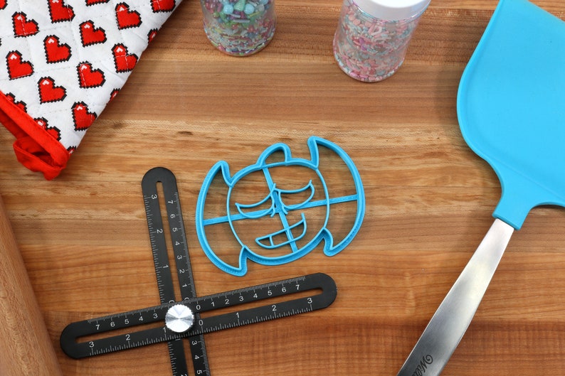 May include: A blue plastic cookie cutter shaped like a cartoon monster with a toothy grin. The cutter is on a wooden surface with a ruler, a rolling pin, and a blue spatula.
