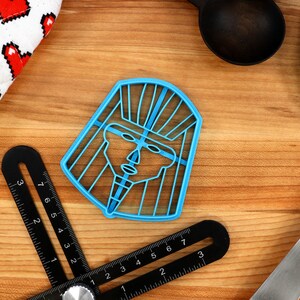 May include: Blue plastic cookie cutter shaped like a stylized human face with a headdress. The cutter is on a wooden surface.