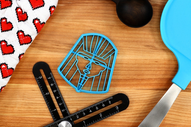 May include: Blue plastic cookie cutter shaped like a stylized human face with a headdress. The cutter is on a wooden surface.