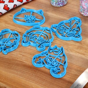 May include: Five blue plastic cookie cutters shaped like cartoon characters. The cutters are on a wooden surface.