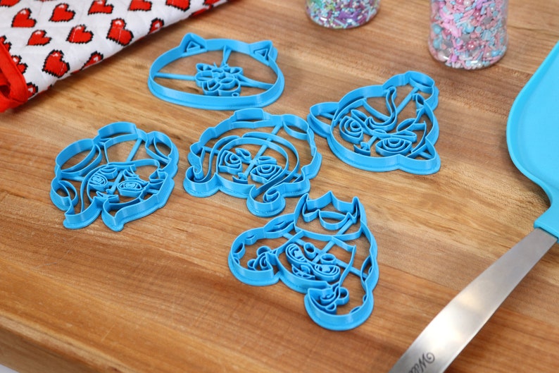 May include: Five blue plastic cookie cutters shaped like cartoon characters. The cutters are on a wooden surface.