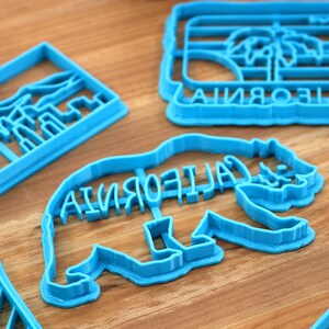 May include: Four blue plastic cookie cutters shaped like California landmarks. The cutters are on a wooden surface. The cutters are shaped like a palm tree with the word "CALIFORNIA" underneath, a bear with the word "CALIFORNIA" on its back, a mountain range, and a striped design.