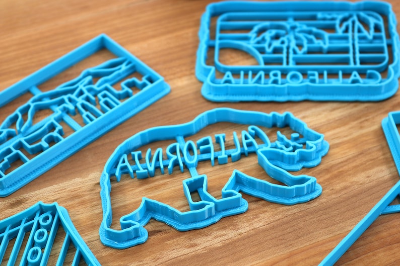 May include: Four blue plastic cookie cutters shaped like California landmarks. The cutters are on a wooden surface. The cutters are shaped like a palm tree with the word "CALIFORNIA" underneath, a bear with the word "CALIFORNIA" on its back, a mountain range, and a striped design.