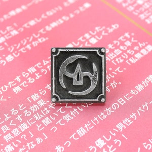 May include: A silver-colored metal pin with a square design. The pin features a stylized symbol with a central point and three curved lines. The pin is on a pink surface.