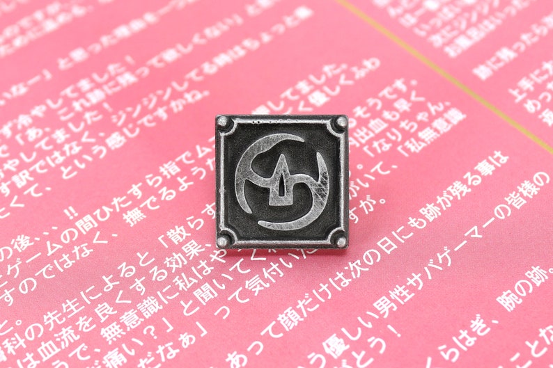 May include: A silver-colored metal pin with a square design. The pin features a stylized symbol with a central point and three curved lines. The pin is on a pink surface.